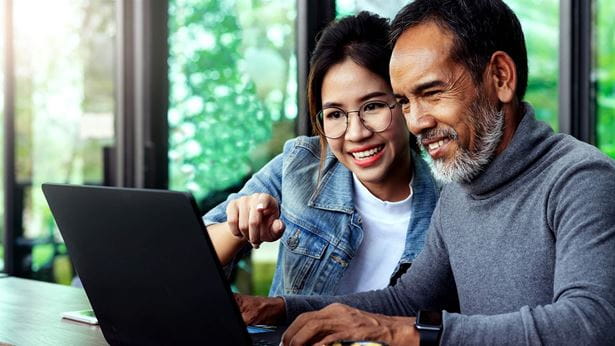 Attractive mature asian man with white stylish short beard looking at laptop computer with teenage eye glasses hipster woman in cafe. Teaching internet online or wifi technology in older man concept.