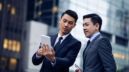 Two asian businessmen talking outdoors. One man is holding his digital tablet and looking at it. Office buildings in back, defocused.