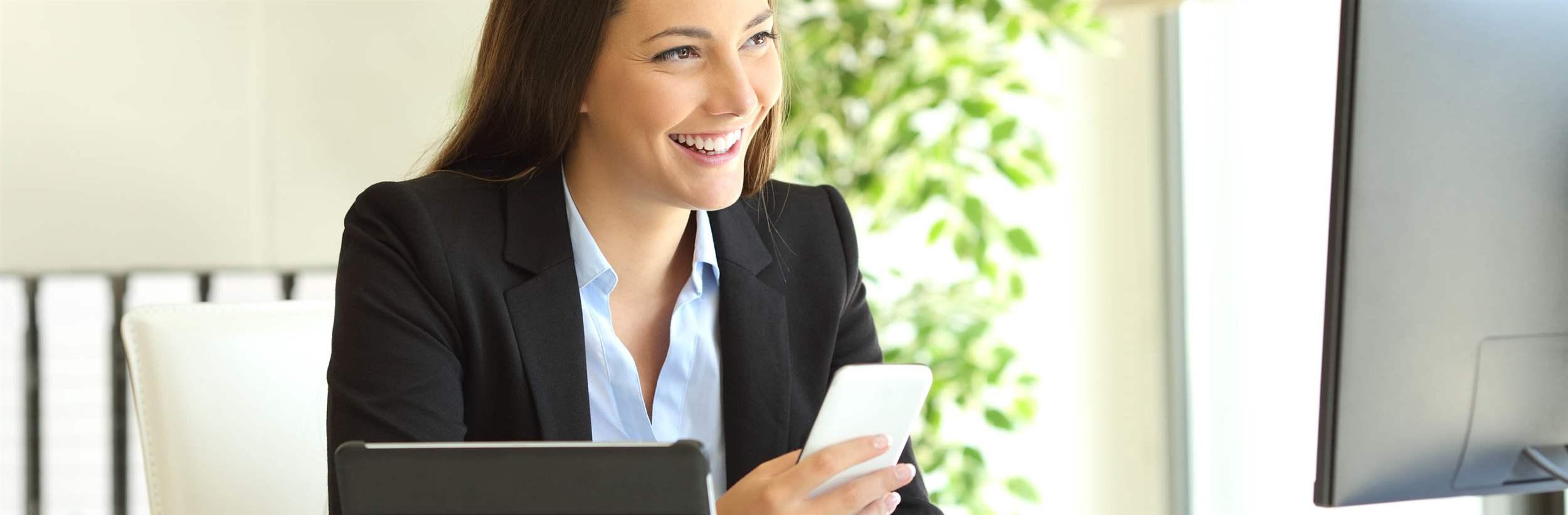 Happy businesswoman working on line with multiple devices and watching a desktop computer at office