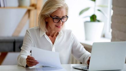 Aged businesswoman typing on pc holds paper prepare report analyzing work results feel satisfied do paperwork use business app online software for data analysis, financier manage firm finances concept