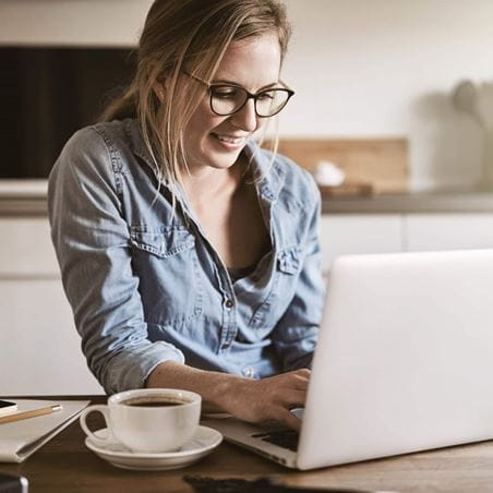 Smiling young woman working on her small business with a laptop while sitting at her kitchen table at home